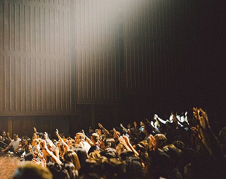 Gruppe junger Menschen steht in Hörsaal vor Bühne, einige von ihnen heben die Hände