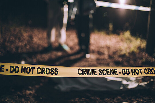 Darstellung eines Tatorts: Im Vordergrund ist ein gelbes Absperrband mit der Aufschrift "Crime Scene - Do not cross" zu sehen. Im Hintergrund sieht man zwei verschwommene Personen. Eine davon trägt einen weißen Schutzanzug der Spurensicherung.
